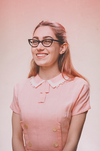 A 1950's styled young woman in a vintage pink dress and cats eye glasses.  Pink background.