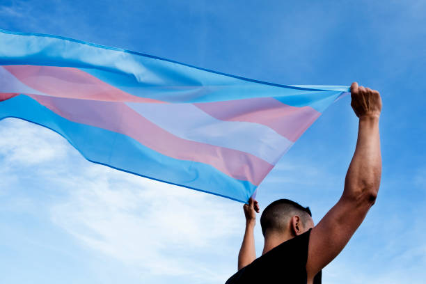 young person with a transgender pride flag a young caucasian person, seen from behind, holding a transgender pride flag over his or her head against the blue sky transgender person stock pictures, royalty-free photos & images
