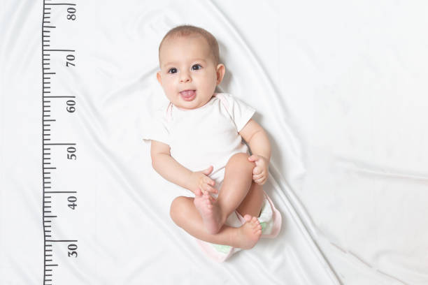 A five month baby in white clothes lying on a bed on which a measuring ruler for growth is drawn. shows the tongue from her mouth and teased stock photo