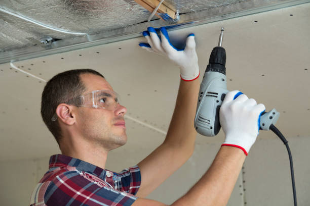 istock Young man in goggles fixing drywall suspended ceiling to metal frame using electrical screwdriver on ceiling insulated with shiny aluminum foil. Renovation, construction, do it yourself concept. 1155293528