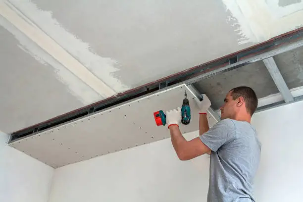 Photo of Young man in usual clothing and work gloves fixing drywall suspended ceiling to metal frame using electrical screwdriver on ceiling insulated with shiny aluminum foil. DIY, do it yourself concept.