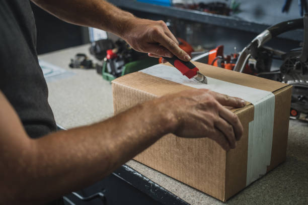 Man opening a box with a knife Hands of a man seen opening a brown cardboard box using a red utility knife. Service bench in the background. Concept of new parts in service. craft knife stock pictures, royalty-free photos & images