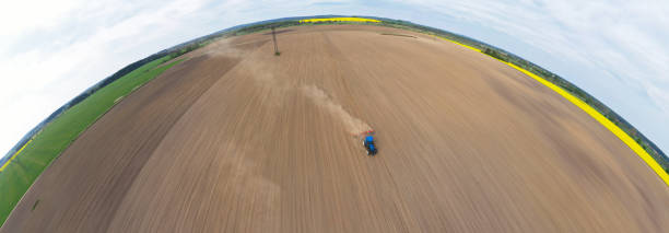 istock Aerial wide curved panoramic view on blue tractor pulling a plow, preparing a soil for seed sowing, tractor making dirt cloud. Landmark on meadow and rapeseed field 1155303526
