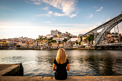 A Portuguese woman admires of the blue sky and soft clouds at sunset displaying Vila Nova de Gaia's colorful City-line from Porto. The double decked metal arched bridge, Don Luís I bridge, overlapping the Douro river, is displayed to the right.