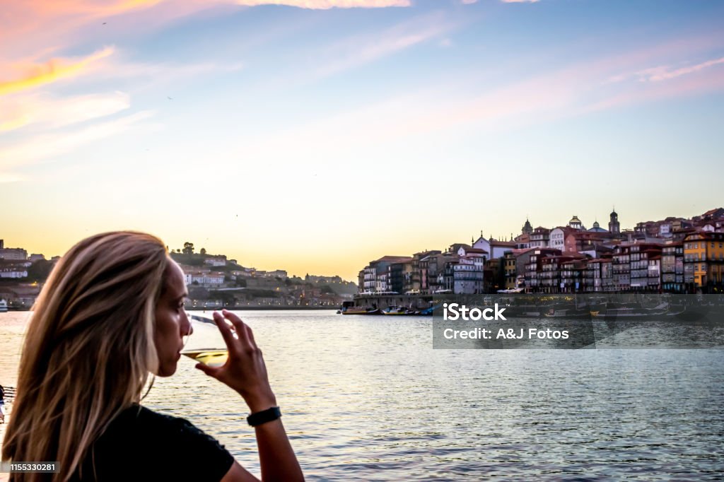 A woman admires the sunset over the Douro river illuminating Porto. A Portuguese woman savors a glass of white wine in admiration of the cotton candy colored sky at sunset displaying it's colorful City-line from Porto. Porto - Portugal Stock Photo