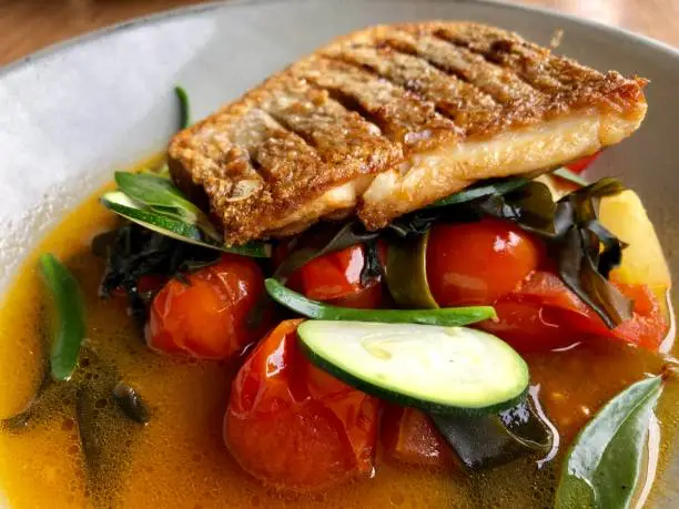 Aerial closeup of a piece of fresh sauted Australian Snapper Fish on a grey ceramic plate with cooked tomatoes and sliced zucchini with green native succulent leaf garnish