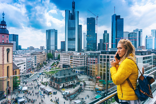Young woman with yellow jacket talking on a smart phone in front of skyline of modern Frankfurt am Main, Germany