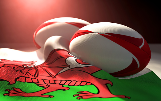 Two regular rugby balls with color design elements resting on a draped wales flag on an isolated dark studio background - 3D render