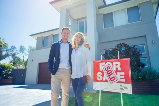 Couple standing in front of their new home with a for sale sign saying sold. Couple standing in front of their new home with a for sale sign saying sold. They are both wearing casual clothes and embracing. The house is contemporary with a driveway, balcony and a green lawn. They are happy and smiling looking at camera. Back lit with lens flare. Copy space in front of photos stock pictures, royalty-free photos & images