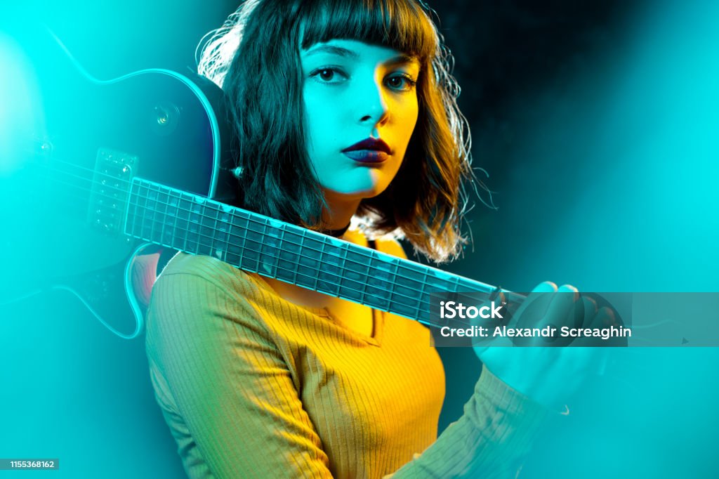Beautiful young hipster woman with curly hair with red guitar in neon lights. Rock musician is playing electrical guitar. 90s style concept. Beautiful young hipster woman with curly hair with red guitar in neon lights. Rock musician is playing electrical guitar. 90s style concept Musician Stock Photo