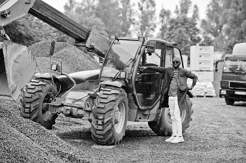 Stylish african american man in hat and sunglasses posed outdoor in rain against tractor with a bucket.