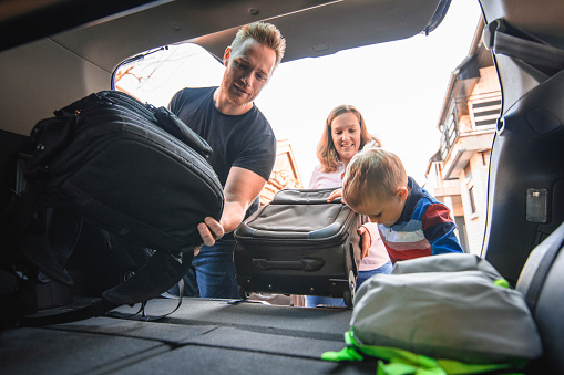 Shot of family with one son packing backpacks and suitcases into car trunk. They are happy and laughing.