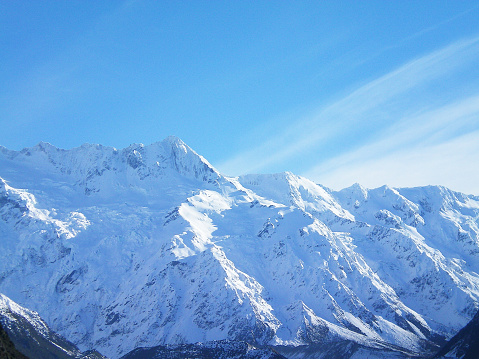 The jagged peaks of snow-covered mountains stand against a blue sky with whispy clouds. Winter sunshine falls on the bright white snow, but the snow in the shadows has tinges of blue.