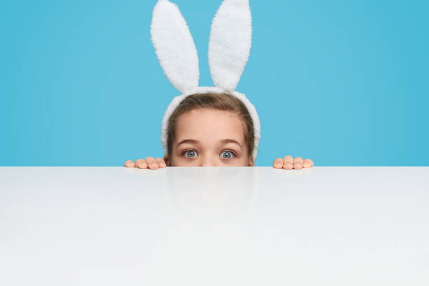 istock Playful girl in bunny ears hiding behind table 1155391428