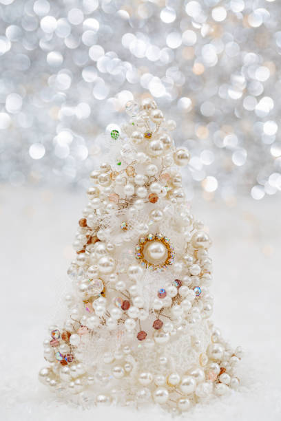 Glamorous white artificial Christmas tree with pearls and beads on the snow next to a beautiful blurred bokeh background and glowing garland.  Shallow depth of field.Toned image. Copy space. Closeup