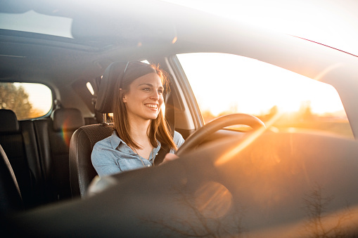 Young woman in a car. She is driving, smiling and looking out. Sun is shining from back on a sunny spring afternoon.