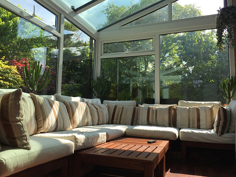 Stock photo of white UPVC conservatory windows with double glazing and glass roof with a beautiful garden view of bonsai trees and blue sky. This conservatory has a sofa with beige and cream cushions, used as inside outside living and dining sun lounge in England, UK