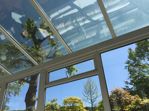 Stock photo of rectangular white UPVC conservatory with glass roof of self-cleaning glass window panes tinted to keep conservatory cool in hot summer weather and not overheating, double-glazed windows with views of landscaped garden trees and sunny blue sky