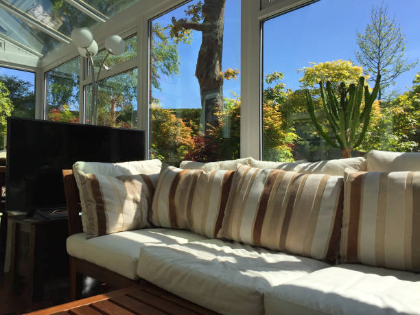 istock Image of rectangular white UPVC conservatory with green garden trees / houseplants, lean-to glass room sunny garden views, double-glazed windows and glass roof with self-cleaning window panes, L-shaped corner sofa settle conservatory furniture, table, TV 1155396493
