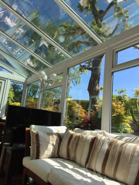 istock Image of rectangular white UPVC conservatory with green garden trees / houseplants, lean-to glass room sunny garden views, double-glazed windows and glass roof with self-cleaning window panes, L-shaped corner sofa settle conservatory furniture, table, TV 1155396495