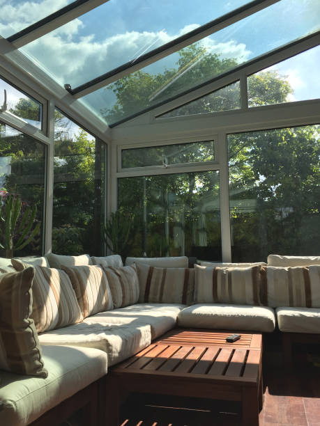 istock Image of rectangular white UPVC conservatory lean-to glass room with sunny garden views, large double-glazed windows and glass roof with self-cleaning panes / L-shaped corner sofa settle conservatory furniture, coffee table and television TV remote contro 1155396509
