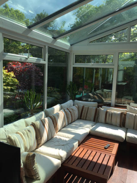 istock Image of rectangular white UPVC conservatory lean-to glass room with sunny garden views, large double-glazed windows and glass roof with self-cleaning panes / L-shaped corner sofa settle conservatory furniture, coffee table and television TV remote contro 1155396539