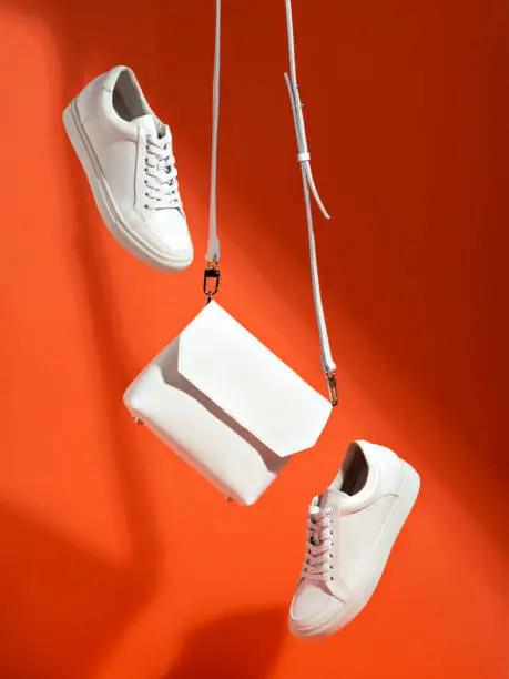 Levitating female white sneakers and small stylish cross body bag on an orange backdrop. Trendy handbag and sport shoes. Summer sale, style or creative outfit concept. Copy space for text.