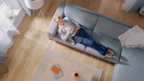 istock Young Man is Lying on a Sofa, Working or Sketcing on a Tablet Computer. Cozy Living Room with Modern Interior, Grey Sofa and Wooden Flooring. Top View Camera Shot. 1155399830