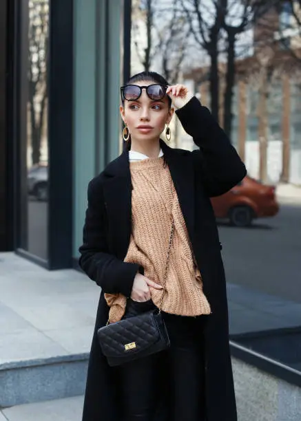 Beautiful girl in sunglasses. A model in a stylish black long coat on a street. Harmoniously clothes similar. Street style . Women's fashion. A girl with a bag over her shoulder.