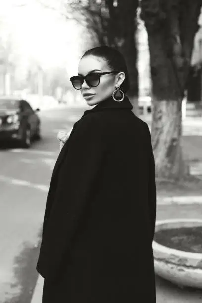 Portrait of a very beautiful girl on a street. Stylish portrait of a pretty girl in a black long coat. hair gathered in a ponytail. The girl on a pedestrian crossing. Black and white photo.