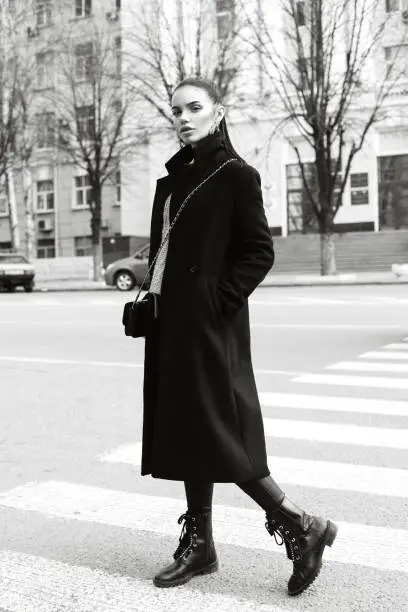 Portrait of a very beautiful girl on a city street. Stylish portrait of a pretty girl in a black long coat. hair gathered in a ponytail. The girl on a pedestrian crossing. Black and white photo.