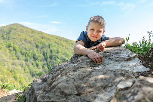 A boy traveler climbed to the top of a cliff surrounded by a green mountains with forest.