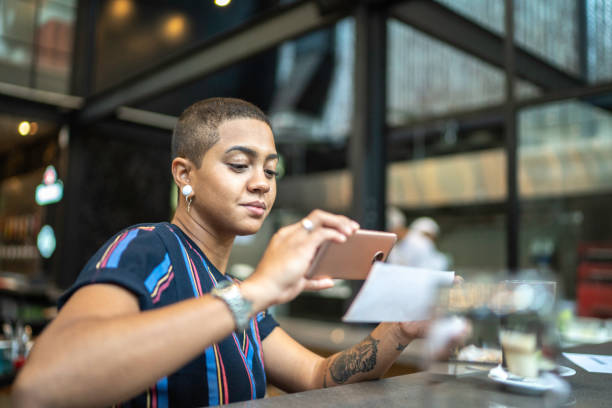 mujer joven depositando cheque por teléfono en el café - dispositivo de información móvil fotos fotografías e imágenes de stock