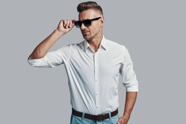 istock Modern macho. Handsome young man smiling and adjusting sunglasses while standing against grey background 1155429188