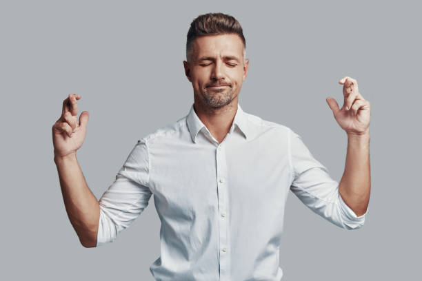 Hopeful. Good looking young man keeping fingers crossed while standing against grey background