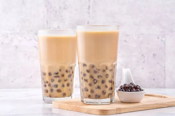 Photo of Popular Taiwan drink - Bubble milk tea with tapioca pearl ball in drinking glass on marble white table wooden tray background, close up, copy space