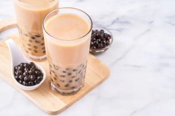 Photo of Popular Taiwan drink - Bubble milk tea with tapioca pearl ball in drinking glass on marble white table wooden tray background, close up, copy space