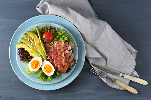 Salad with avocado and bacon. In a deep plate lettuce leaves, fried bacon, avocado slices, boiled egg, tomato, pine nuts. In the frame napkin and cutlery. Dark background. Close-up. View from above.