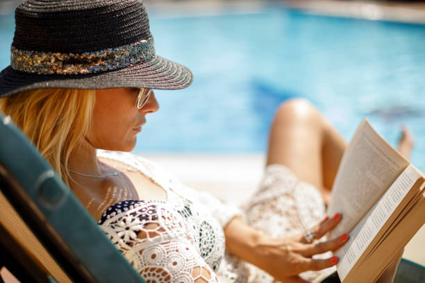femme avec le chapeau et les lunettes de soleil utilisant la lecture d’un livre dans son arrière-cour - rebord de piscine photos et images de collection
