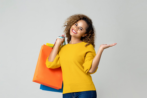 Young smiling African American woman holding colorful shopping bags with another hand open on light gray background