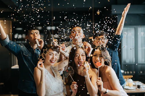 Group of cheerful young Asian man and woman holding props and celebrating with confetti during party