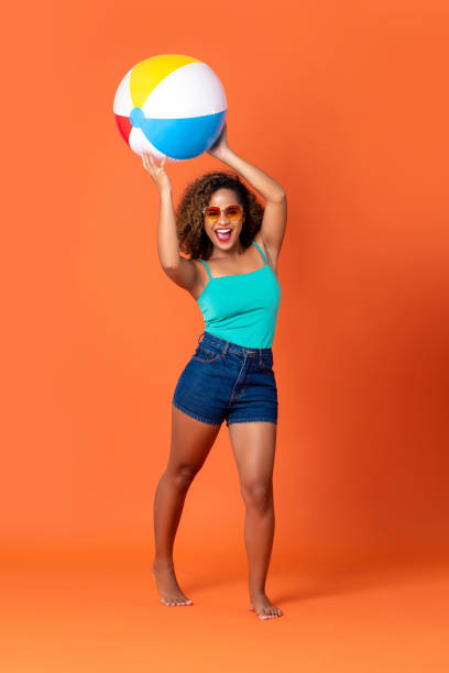 Pretty smiling African American woman in summer concept Pretty smiling African American woman in casual summer clothes with colorful beach ball on orange background beach ball beach summer ball stock pictures, royalty-free photos & images