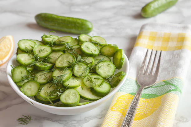 Quick and easy thinly sliced cucumbers and dill salad on the plate with whole cucumbers, lemon and fork on the table-napkin in white colors in the kitchen. Healthy habits homemade veggie tables concept - image