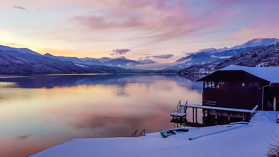 Beautiful view on Millstätter lake in Austria. The lake is surrounded by Alps. Mountains are covered with snow. The sky is exploding with pink and orange. Stunning sunset. Little cottage on the side.