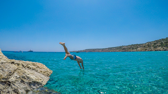 A young athletic-build man jumping into the sea in Cape Greco, Cyprus. Water is crystal clear, shimmering with many shades of blue. Jump of joy. Man is wearing blue swimsuit.