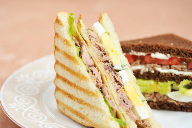On the plate are two triangular-shaped sandwiches. One sandwich of white bread, the other of dark. Filling of tuna sandwiches, hard boiled eggs and vegetables. Light background. Close-up.