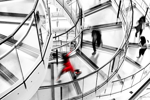 High angle image depicting an abstract view of a group of people running down a modern spiral staircase in the city. The running is depicted as motion blur, which makes it looks as though the people are moving extremely fast. Everything in the image is desaturated except one female who is dressed entirely in red. Room for copy space. ***IMAGE SHOT IN CITY HALL, LONDON, UK, A PUBLICLY OWNED BUILDING FREELY ACCESSIBLE TO THE PUBLIC. PLEASE NOTE THERE NO FEES OR TICKETS ARE REQUIRED TO ENTER, AND THERE ARE NO PHOTOGRAPHIC RESTRICTIONS***