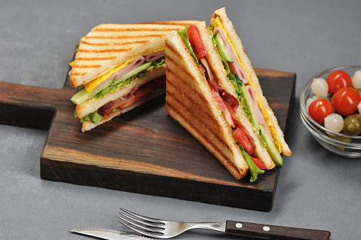 Classic club sandwich with ham and bacon on a wooden board. Next cup is a mixture of pickled miniature onions, gherkins, tomatoes. Gray background. Close-up. Macro photography.
