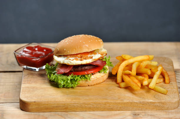 istock Hamburger on a light wooden board. The burger includes bacon, fried egg and dried onions. Next to the burger are french fries and ketchup cups. 1155511632
