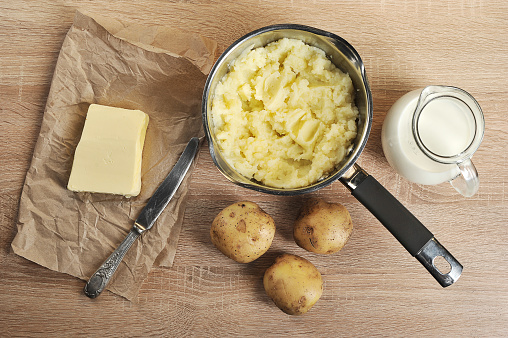 Set for the preparation of mashed potatoes. In a saucepan boiled potatoes, a jug with milk, butter. Light wooden background. Close-up. View from above.